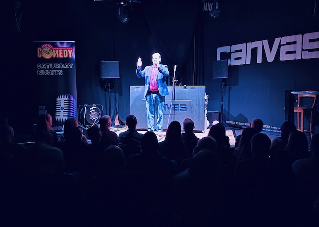 Stand-up comedian performing on stage at Canvas venue with Jaggers Comedy Club branding and engaged audience